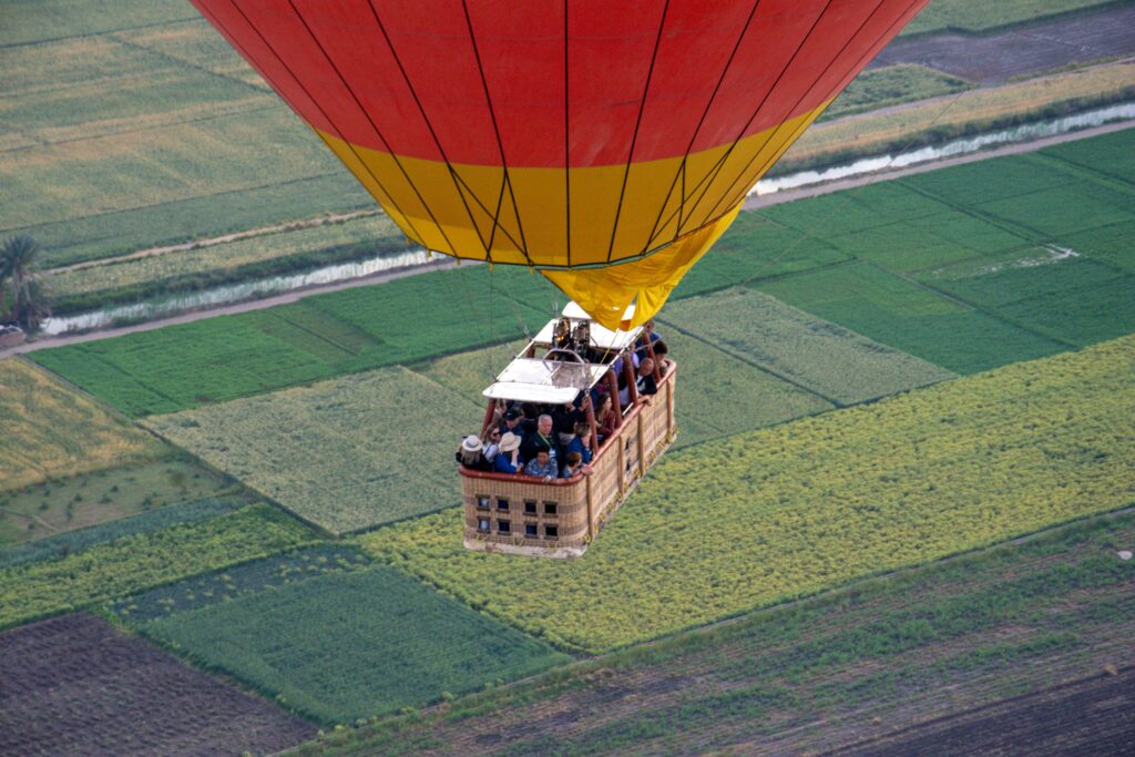 Tourists in a hot air balloon's basket flying over a green field across the Nile